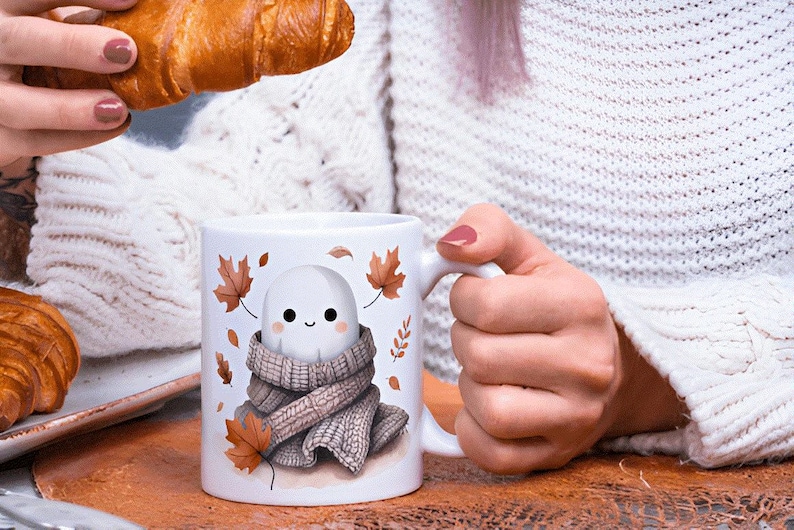 May include: White ceramic mug with a cute ghost character in a scarf and autumn leaves. The mug is held by a person. A croissant is held in the other hand. The mug is on a wooden surface. The mug has a handle.