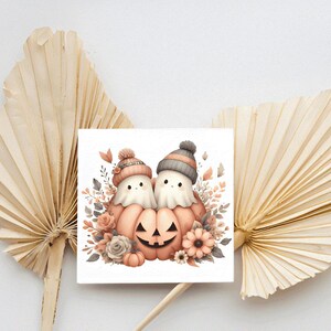 May include: A square print depicting two ghost characters in knitted hats, positioned around a carved pumpkin. The illustration uses soft watercolour tones of peach, grey, and cream, with floral accents. The print is set against a neutral background.