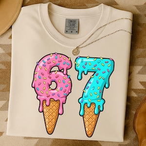 Op de afbeelding: Een crèmekleurig T-shirt met het nummer "67" ontworpen als ijsjes. De "6" is roze met hagelslag en de "7" is blauw met hagelslag. Het shirt heeft een Comfort Colors label en een gouden ketting.