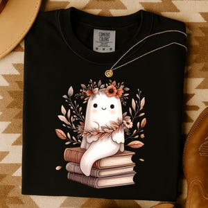 May include: Black t-shirt with a cartoon ghost wearing a floral crown, perched on books. The ghost has a smiling face. The shirt has a Comfort Colors tag. A gold necklace is draped across the shirt.