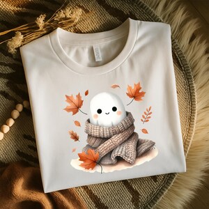 May include: A white t-shirt with a cartoon ghost wearing a knitted scarf, surrounded by orange and brown autumn leaves. The shirt is displayed on a textured, neutral-toned surface. The design evokes a cosy autumn theme.