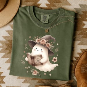 May include: Green Comfort Colors t-shirt with a cute ghost illustration. The ghost wears a witch's hat decorated with flowers and reads a book. Floral and star details complete the design, offering a charming, spooky aesthetic.