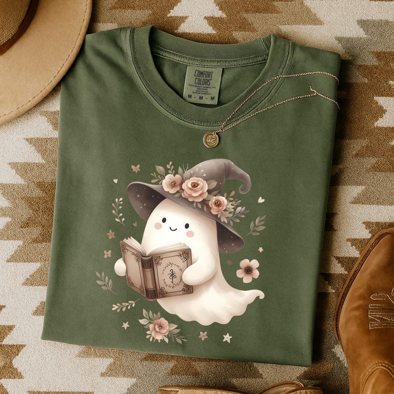 May include: Green Comfort Colors t-shirt with a cute ghost illustration. The ghost wears a witch's hat decorated with flowers and reads a book. Floral and star details complete the design, offering a charming, spooky aesthetic.