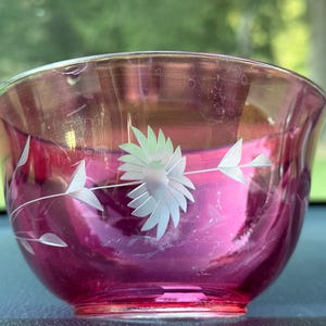 May include: A ruby-colored glass bowl with a floral design etched into the surface. The bowl features a large flower and leaf pattern in white. The bowl is likely vintage and is a decorative item.