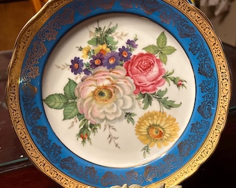 Könnte beinhalten: Ein dekorativer Porzellanteller mit floralem Design. Der Teller zeigt einen zentralen Blumenstrauß aus rosa, weißen, gelben und lila Blumen. Ein blauer Rand mit goldenen Verzierungen umgibt das Blumenarrangement. Der Rand ist mit Gold verziert.