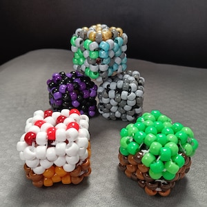 May include: Five colorful beaded dice, each with a different color pattern. The dice are made of small plastic beads and are about 2 inches in diameter. The colors include green, purple, white, gray, and gold.