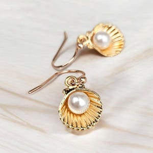 May include: Gold-tone seashell earrings, each featuring a white pearl. The earrings have a hook fastening. The seashell design is detailed with textured lines. The earrings are set against a white background.
