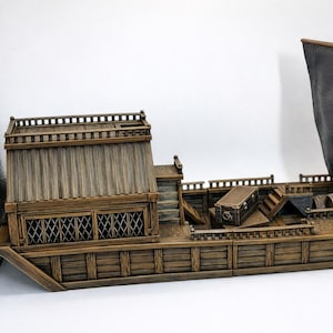 Puede incluir: Maqueta de madera detallada de un barco con una gran vela. El barco presenta una cabina, detalles de cubierta y una vela gris oscuro. El modelo está pintado en tonos marrones y grises, dándole un aspecto envejecido.