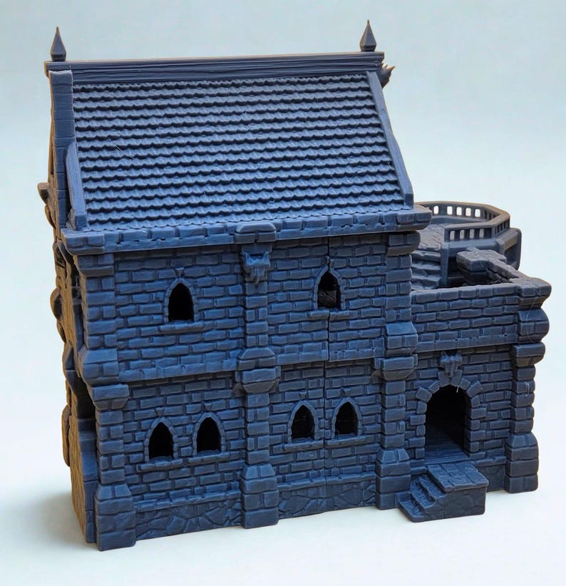 May include: A dark blue, 3D-printed miniature building featuring a two-story stone structure with a tiled roof, arched windows, and a side terrace with stairs. The exterior walls display a detailed brick pattern, and the roof has pointed finials.