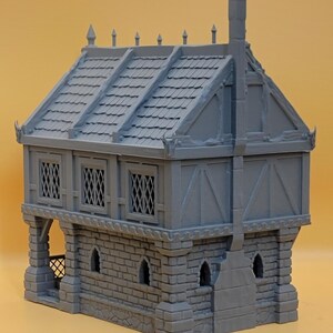 May include: A gray miniature building model featuring a stone-textured ground floor with arched windows and a timber-framed upper level with lattice windows. The structure includes a shingled roof with decorative finials and a prominent chimney.