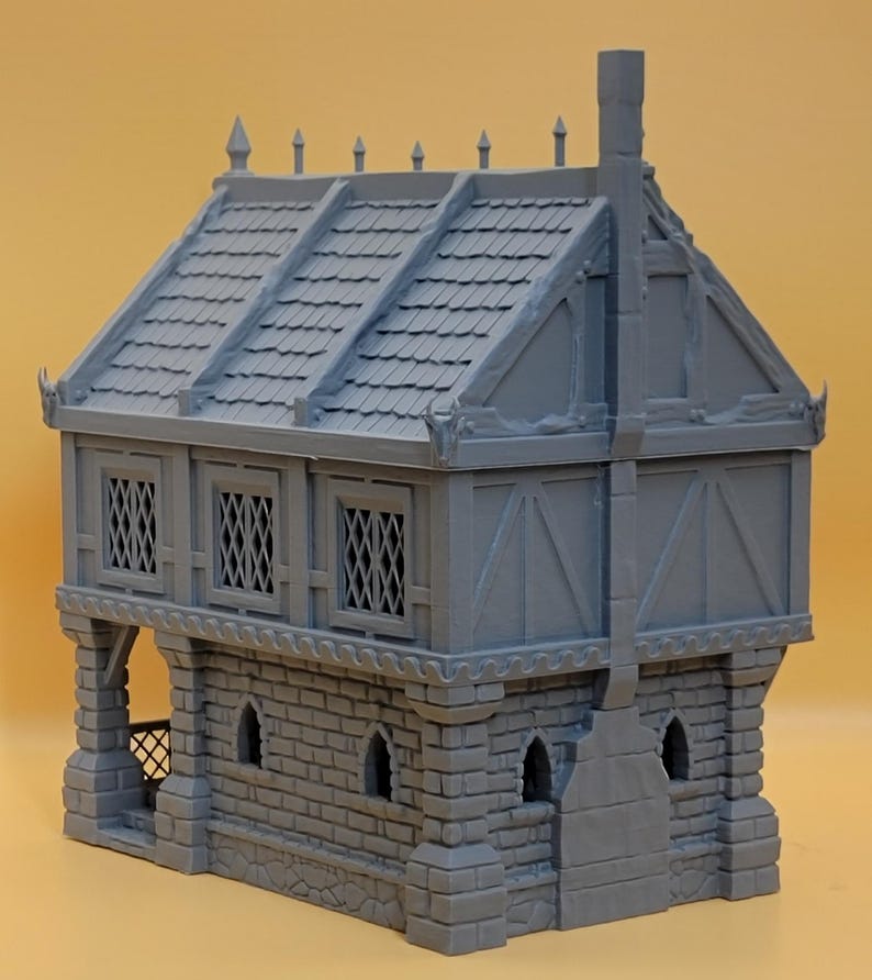 May include: A gray miniature building model featuring a stone-textured ground floor with arched windows and a timber-framed upper level with lattice windows. The structure includes a shingled roof with decorative finials and a prominent chimney.