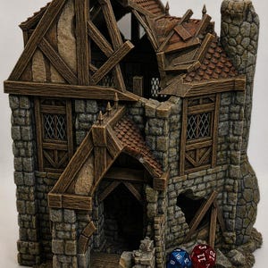 Op de afbeelding: Een gedetailleerde miniatuur van een verweerd stenen gebouw met een beschadigd dak en houten balken. De structuur heeft een schoorsteen en er zijn twee dobbelstenen zichtbaar. Het gebouw is waarschijnlijk voor tabletop gaming.