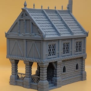 May include: A gray 3D printed miniature building featuring a stone-textured ground floor with arched windows and a timber-framed upper level with lattice windows. The structure includes a covered porch area with stone pillars and a tiled roof.