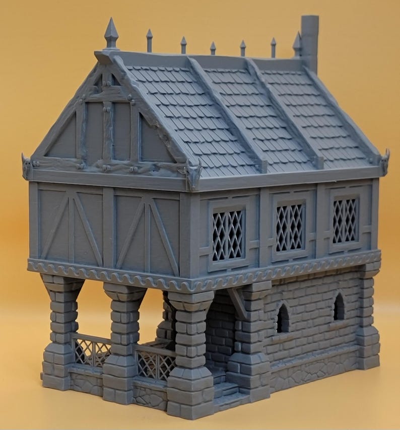 May include: A gray 3D printed miniature building featuring a stone-textured ground floor with arched windows and a timber-framed upper level with lattice windows. The structure includes a covered porch area with stone pillars and a tiled roof.