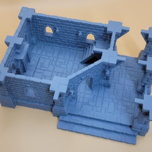 May include: A light blue, 3D-printed miniature stone building structure featuring a rectangular room with brick-textured walls, arched window openings, a small staircase leading to an upper level, and an open-air courtyard area with stone flooring.
