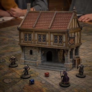 May include: A miniature two-story building with a brown shingled roof, timber-framed upper walls, and a stone-textured ground floor. A hanging sign is attached to the side. Four small character figurines and two dice are placed on the grid surface.