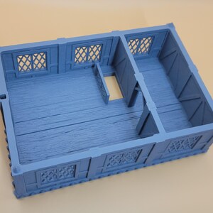 May include: A light blue, rectangular, miniature building structure featuring a divided interior floor plan with wood grain texture, exterior walls with lattice-style windows, and a single open doorway connecting the two separate rooms.
