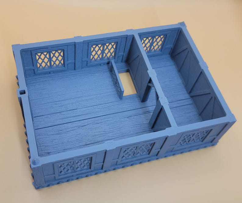 May include: A light blue, rectangular, miniature building structure featuring a divided interior floor plan with wood grain texture, exterior walls with lattice-style windows, and a single open doorway connecting the two separate rooms.