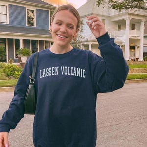 May include: A navy blue sweatshirt with the text "LASSEN VOLCANIC" in white block letters. The sweatshirt is worn by a person outdoors, with a black handbag over their shoulder. The background includes houses and a street.