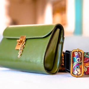 Puede incluir: Un bolso de mano de cuero verde con un cierre y cadena dorados, junto a una pulsera de puño estampada con un marco dorado. La pulsera presenta un diseño abstracto y colorido. El bolso mide unos 25 cm de ancho.