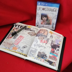 May include: An open notebook with a black cover and white pages filled with colorful stickers and drawings. The notebook is open to a page with a sticker that says "Arcadia Bigfoots" and another sticker that says "Life is Strange". The notebook is lying on a red surface.