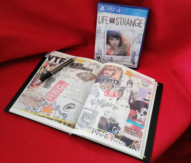 May include: An open notebook with a black cover and white pages filled with colorful stickers and drawings. The notebook is open to a page with a sticker that says "Arcadia Bigfoots" and another sticker that says "Life is Strange". The notebook is lying on a red surface.
