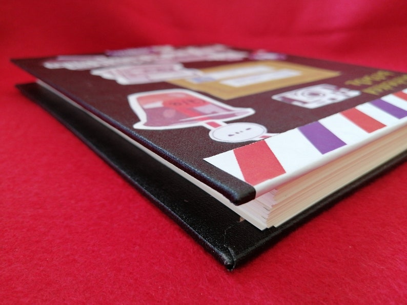May include: A black hardcover journal with a colorful design on the cover. The design features a red bell, a purple and red striped pattern, and the text "Postcards from the World".
