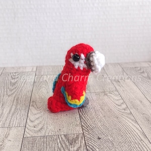 May include: A small, crocheted red parrot with a white beak and black eyes. The parrot has blue and yellow accents on its wings.