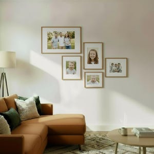 May include: A collection of five framed photographs with light brown wooden frames. The photos feature family portraits and individual portraits. The frames are arranged on a white wall, creating a gallery-style display. The room also includes a brown sectional sofa.