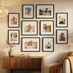 May include: A gallery wall arrangement featuring nine black rectangular picture frames with white matting, displaying various family photographs. The frames are organized in a grid-like layout on a plain wall above a wooden console table.