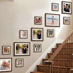 May include: A gallery wall arrangement featuring twelve dark brown wooden picture frames of varying sizes, displayed in a staggered formation along a staircase wall, each containing a different personal photograph with white matting.