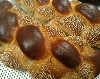 May include: Close-up of a braided loaf of bread. The bread has a golden-brown crust with dark brown, glossy sections. It is generously covered with sesame seeds, creating a textured appearance. A delicious baked good.