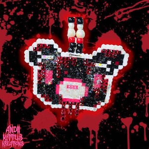 May include: A pixelated bear pendant with a white outline, black and pink details, and red glitter accents. The pendant is attached to a beaded chain with black, white, and red beads. The background is black with red splatter and the text "Andii Kittys Kreations".