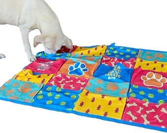 May include: A multi-colored dog play mat with pockets for hiding treats. The mat has paw prints, bones, and tennis balls printed on it. A dog is sniffing the mat.