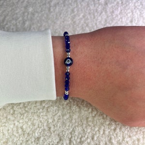 Puede incluir: Una pulsera de cuentas azules con un dije de ojo turco. La pulsera presenta pequeñas cuentas azules facetadas y un dije central con un diseño de ojo azul y blanco. La pulsera se usa en una muñeca, sobre un fondo blanco.