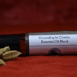 Puede incluir: Un frasco rodillo de mezcla de aceites esenciales marrón con tapa negra, etiquetado como "Grounding 1st Chakra Essential Oil Blend". El frasco descansa sobre una superficie roja junto a varias vainas de cardamomo marrón claro. La etiqueta también incluye las palabras "Oak & Thistle Apothecary".