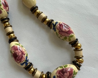 May include: A white ceramic necklace with gold and black beads and floral designs. The flowers are pink, blue, and green.