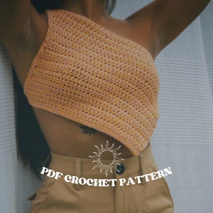 Może przedstawiać: Brzoskwiniowy top z jednym ramieniem, wykonany na szydełku. Top jest ręcznie robiony, ma teksturowy ścieg i unikalny asymetryczny design. Tekst "PDF CROCHET PATTERN" jest wyświetlany na biało, wraz ze słońcem.