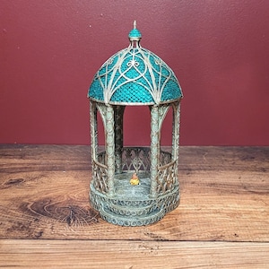 May include: A detailed miniature gazebo with a teal roof and intricate silver metalwork. The gazebo has a domed roof and decorative lattice sides, with a small figure inside. The gazebo sits on a wooden surface.