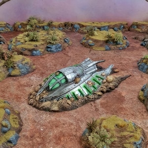 Op de afbeelding: Een zilveren en groen ruimtevaartuigmodel is neergestort op een woestijnlandschap met verschillende kleine heuvels en twee bomen. Het model is omringd door zand en rotsen. De tekst "Printable Scenery" en "www.printablescenery.com" is zichtbaar in de rechterbenedenhoek van de afbeelding.