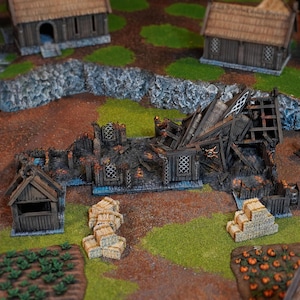Op de afbeelding: Een tabletop-spelset met een verwoest houten gebouw met geblakerde muren en een ingestort dak, omringd door kleine houten structuren, stapels hooibalen, een moestuin en een pompoenveld op een speelmat.