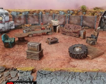 May include: A miniature model of a post-apocalyptic campsite with a couch, chairs, a table, a fire pit, a television, and a tire. The scene is set on a dirt ground with a chain link fence in the background.