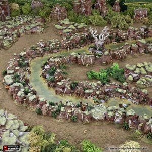 May include: A miniature tabletop gaming terrain set featuring a winding river flowing through a mossy, rocky landscape. The terrain is made of brown, green, and grey materials, and includes a variety of trees and rocks.
