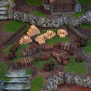 May include: A detailed miniature diorama with wooden crates, barrels, and hay bales. Some crates are filled with red objects. The scene includes a small wooden building and stone walls, set on a grassy terrain.