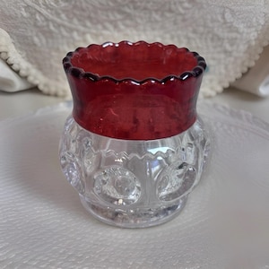 May include: A clear glass vase with a ruby red rim and a scalloped edge. The lower portion of the vase has a pattern of circular designs. The vase is likely vintage and decorative.