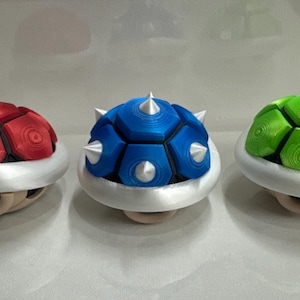 May include: Three 3D printed Super Mario Bros. Koopa Troopa shells. One is red, one is blue, and one is green. Each shell has a white base.