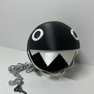 May include: A black and white 3D printed hanging planter shaped like a character with a large open mouth and two white eyes. The planter is attached to a silver chain with a hook.