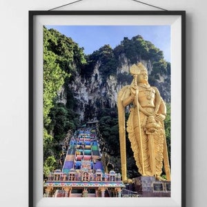 May include: A large golden statue of a Hindu deity stands in front of a colorful staircase leading up to a temple. The statue is holding a staff and is surrounded by lush green foliage.