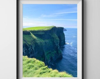 May include: A framed wall art print featuring a landscape photograph of steep, dark green cliffs overlooking a deep blue ocean under a bright, clear sky with thin white clouds. The frame is black with a white mat border and a hanging wire.