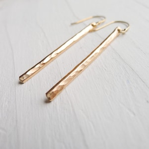 May include: A pair of long, gold-toned earrings with a hammered texture. The earrings are made of a thin, rectangular piece of metal that is slightly curved.
